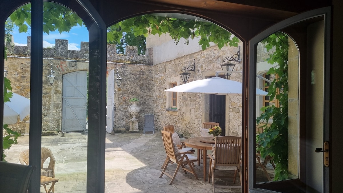 An outdoor courtyard is viewed through large arched doors, framed by green vines overhead. Wooden chairs and a table are arranged on the stone-paved surface. A sun umbrella provides shade, while stone walls with decorative lighting fixtures create a serene atmosphere.