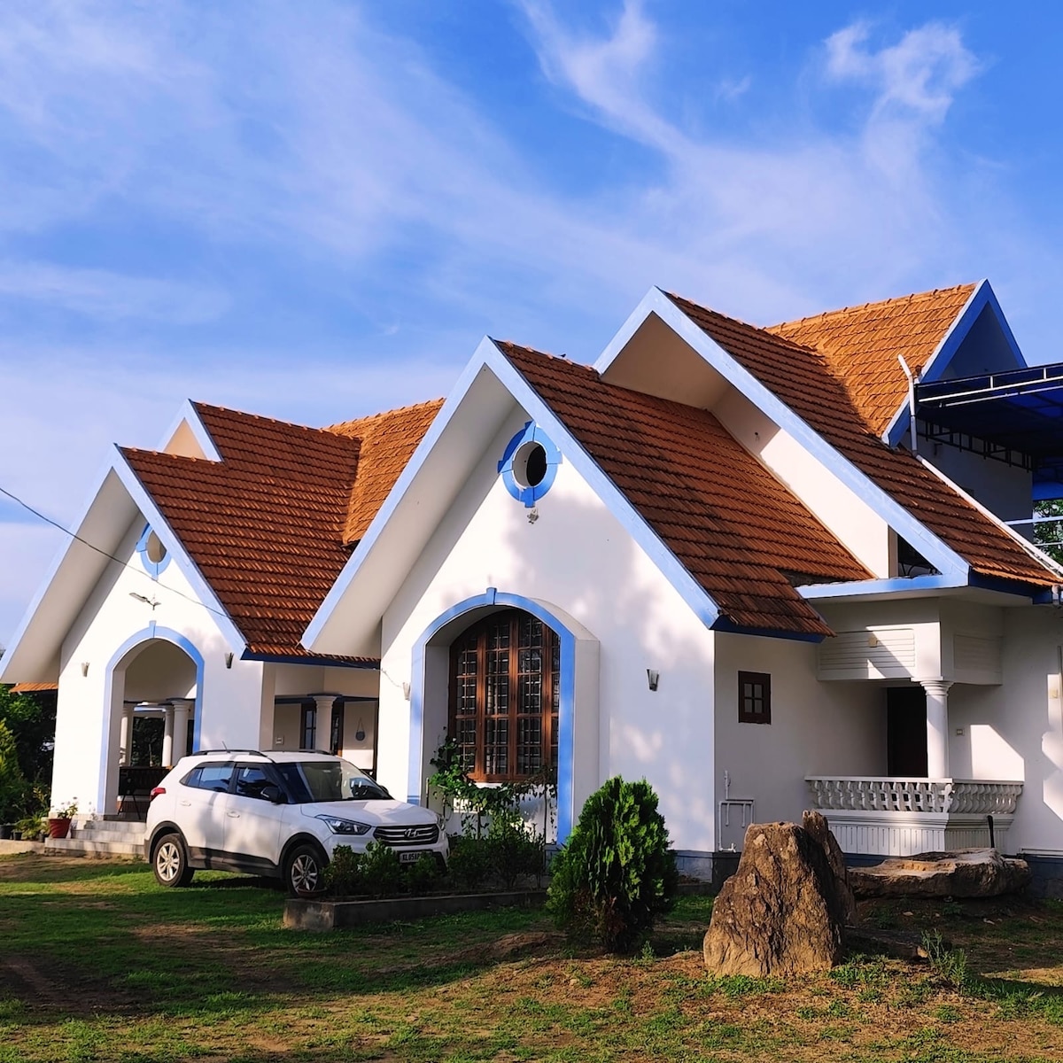 The homestay features a charming exterior with a distinctive roof and classic white walls. Large windows enhance the inviting appearance, while a car is parked in front. Lush greenery surrounds the property, contributing to a serene atmosphere, complemented by clear blue skies.