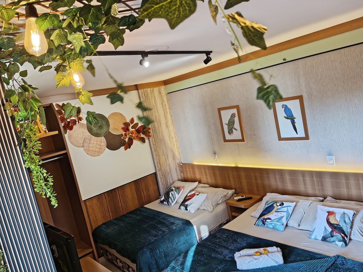 The suite features a bright and airy layout with two spacious beds adorned with colorful bird-themed bedding. Decorative foliage hangs from the ceiling, and wall art pieces of birds are displayed. A wardrobe is visible, providing convenient storage space.