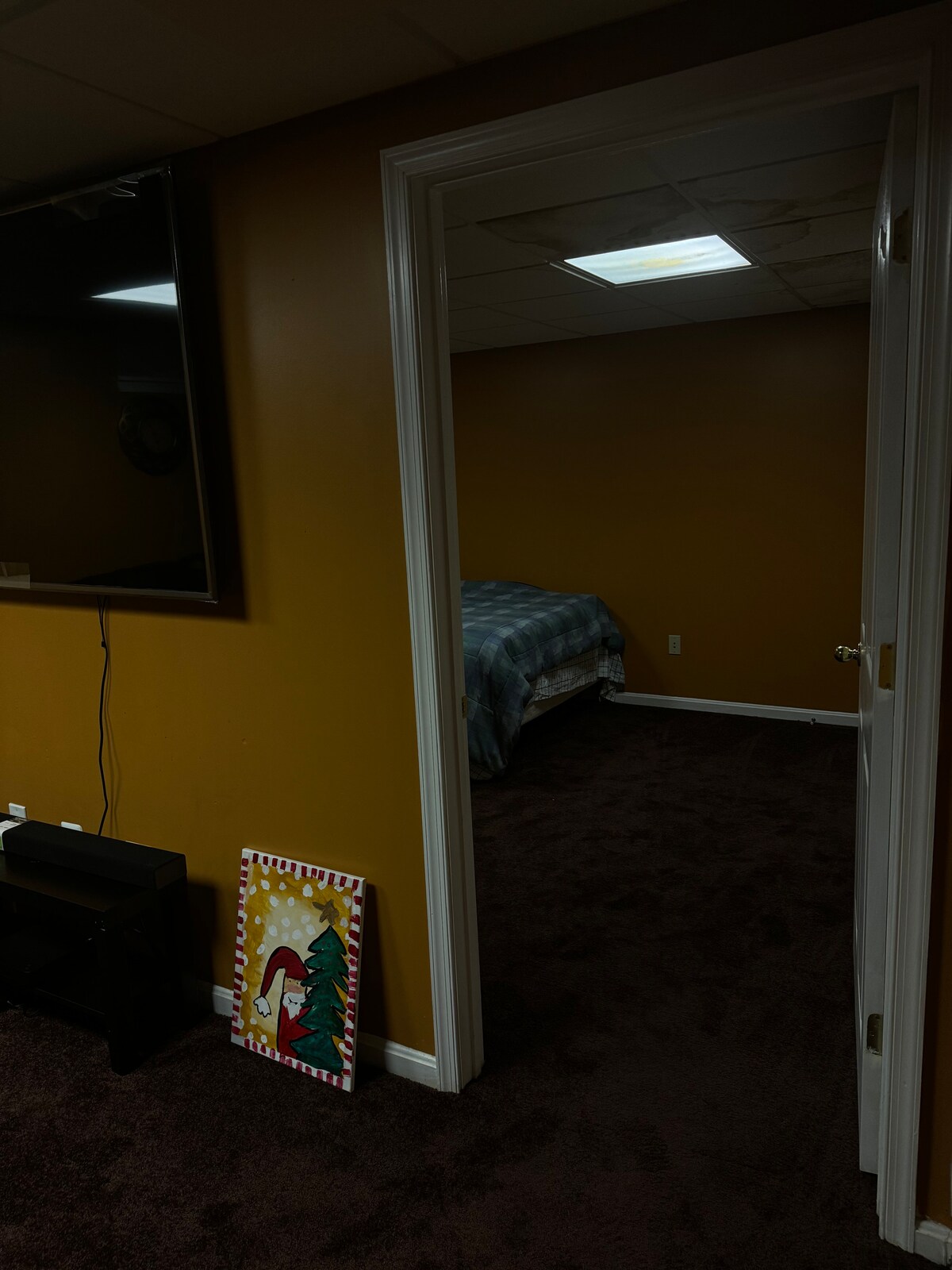 A doorway leads into a spacious bedroom with warm, golden walls and a plush, dark carpet. A cozy bed is partially visible in the background, covered with a blue and gray plaid blanket. A wall-mounted TV is positioned to the left, enhancing the room's comfort.