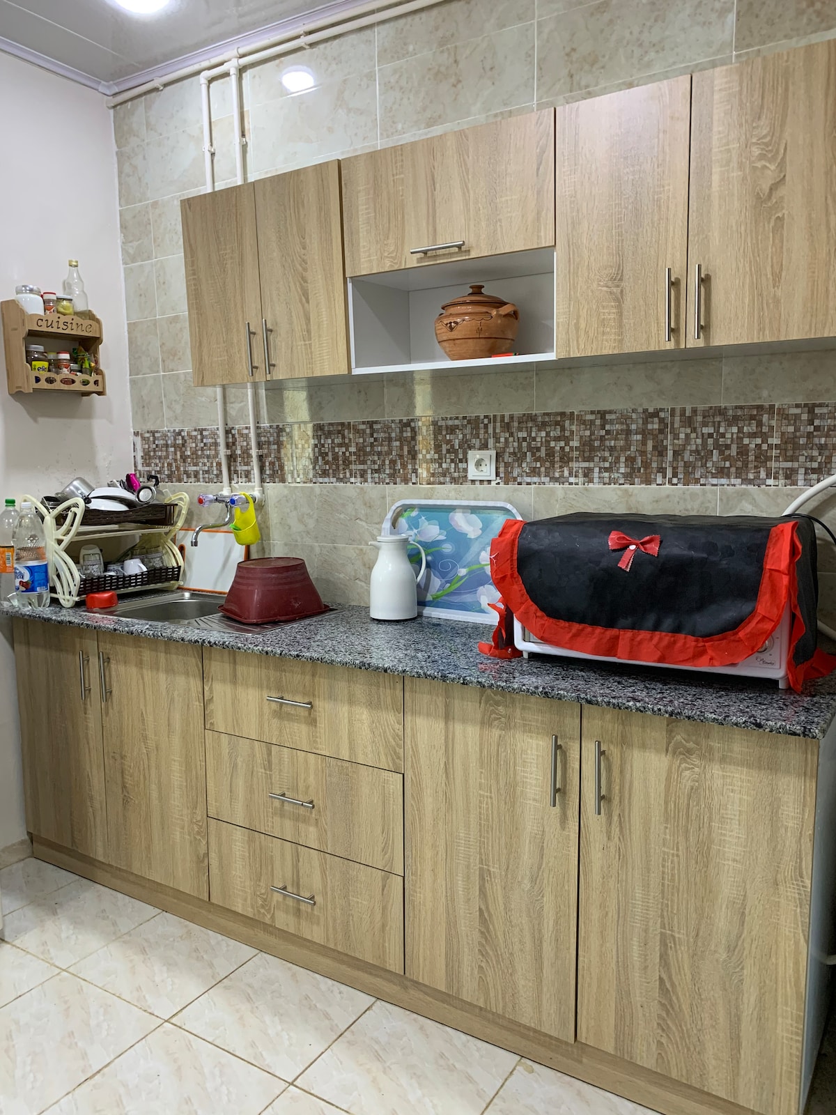 A compact kitchen features wooden cabinetry with a granite countertop. A sink is positioned near colorful kitchen items, including a red container and a white bottle. Above the counter, shelves hold a decorative pot and additional kitchenware, adding functionality to the space.