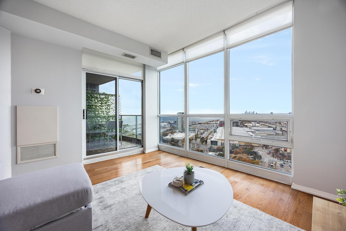 A spacious living area is showcased, featuring large floor-to-ceiling windows that provide ample natural light and picturesque views of the cityscape and Lake Ontario. A light-colored coffee table sits in the center, while a cozy seating arrangement includes a plush couch, inviting relaxation.
