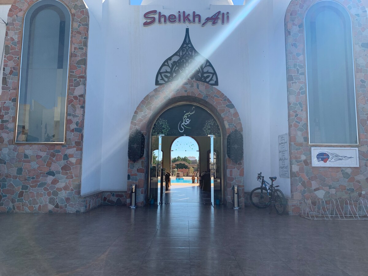 The entrance of Sheikh Ali Dahab Resort is framed by large, arched doors with intricate designs. Stone accents and greenery enhance the exterior. A glimpse of the pool area is visible through the entrance, offering an inviting view of the resort’s surroundings.