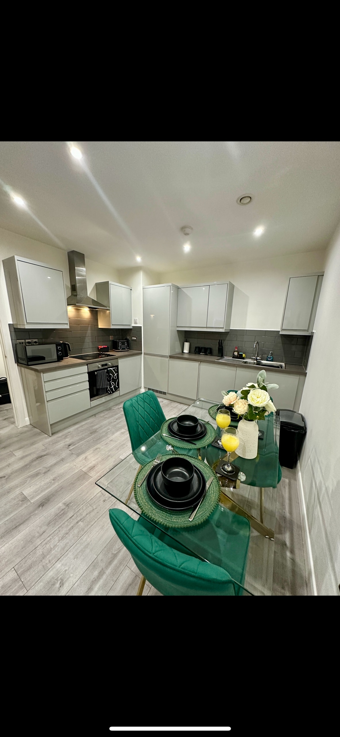 A modern kitchen is featured, equipped with sleek, grey cabinetry and a spacious countertop. A glass dining table is set with plates, glasses, and a floral centerpiece. Green chairs complement the table, enhancing the stylish ambiance. The appliances, including a microwave and oven, are visible in the background.