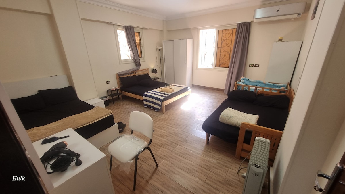 A spacious room is furnished with three beds and a baby bed. A chair and a table are positioned in the corner, while natural light filters through windows adorned with light drapes. Wooden flooring complements the simple decor, creating an open atmosphere.