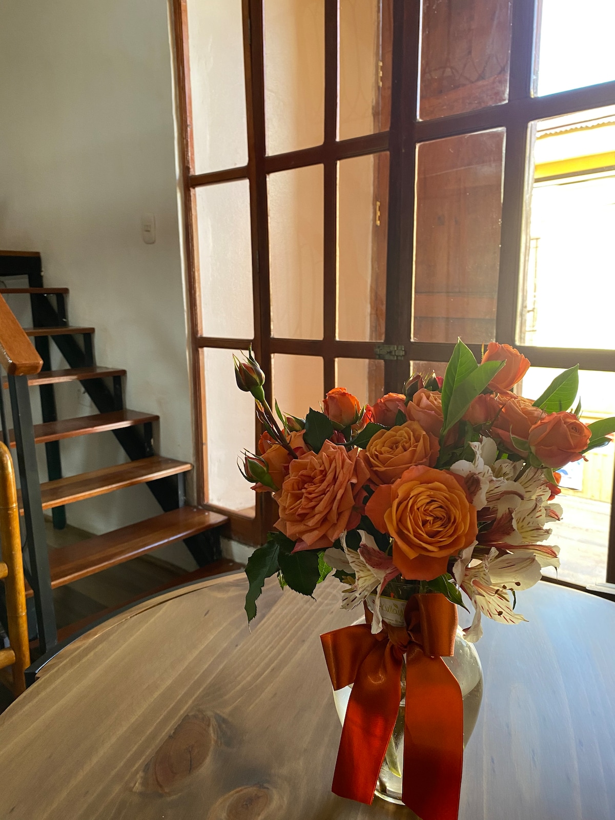 A wooden table is adorned with a vase containing an arrangement of orange roses and white flowers. Natural light filters through large windows, highlighting the wooden staircase in the background.