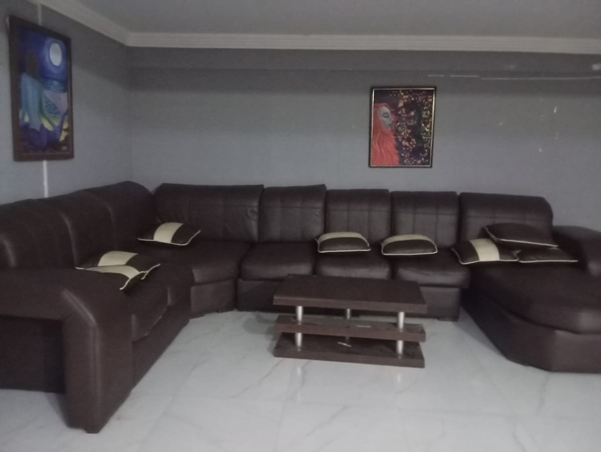 A spacious living area features a large dark leather sectional sofa adorned with decorative pillows. A low wooden coffee table is positioned in front, enhancing the comfortable seating arrangement. Colorful wall art adds visual interest to the neutral grey walls.