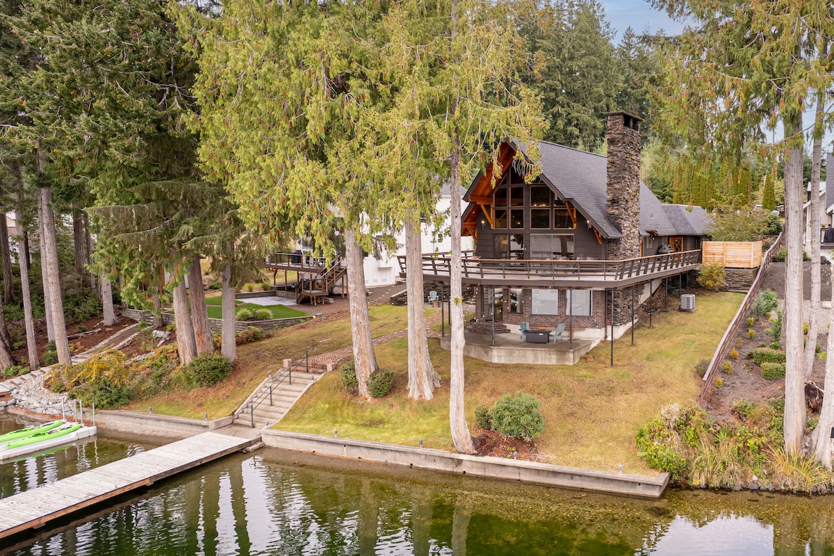 A lakeside A-frame cabin is nestled among trees, featuring a spacious deck that overlooks Mason Lake. The private dock provides access to the water, where kayaks are visible. The surrounding landscape includes manicured lawns and natural greenery, creating a peaceful outdoor environment.