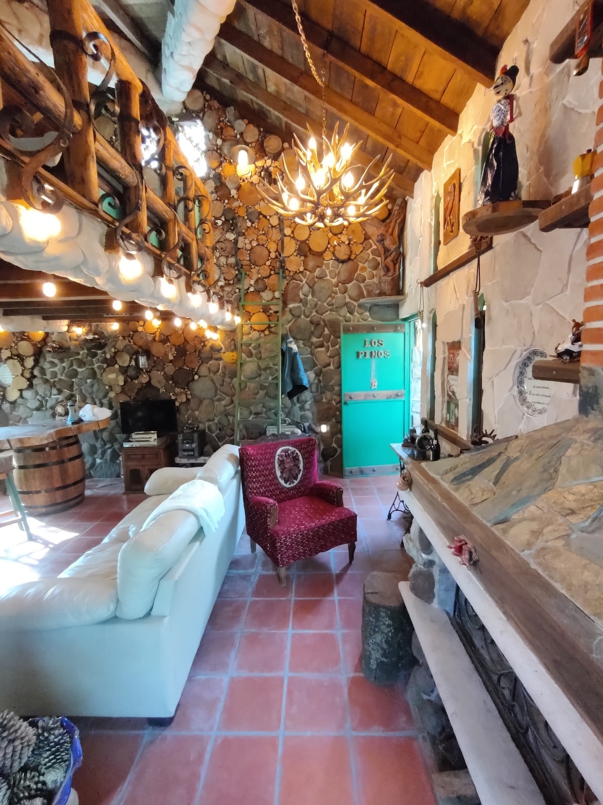 A cozy interior features a mix of rustic charm and artistic elements, highlighted by a high wooden ceiling. A fireplace and a white couch create a warm gathering area, while a red upholstered chair adds a pop of color. Soft lighting from string lights enhances the atmosphere.