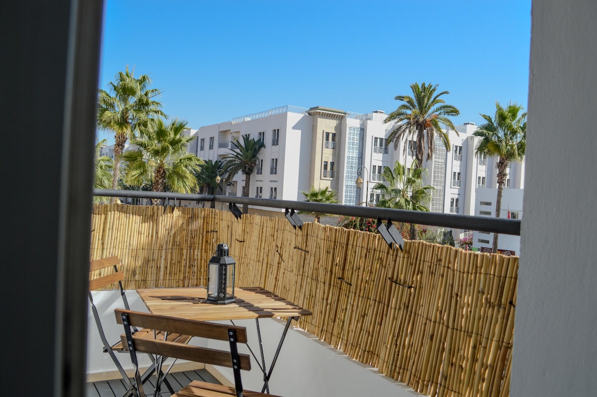 A balcony is showcased, featuring a wooden table and two folding chairs. A bamboo screen provides privacy while palm trees and neighboring buildings are visible in the background, contributing to a relaxed outdoor setting with clear blue skies above.