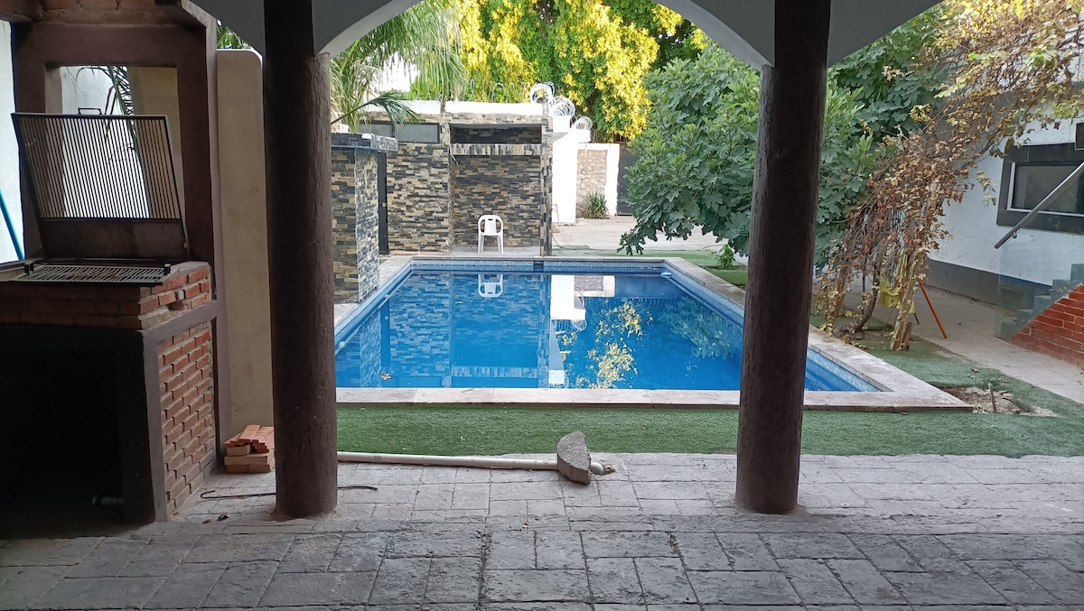A serene view of a swimming pool is framed by arching columns. The pool's clear water reflects the surrounding greenery. A barbecue grill stands nearby, offering a designated space for outdoor cooking and gathering.