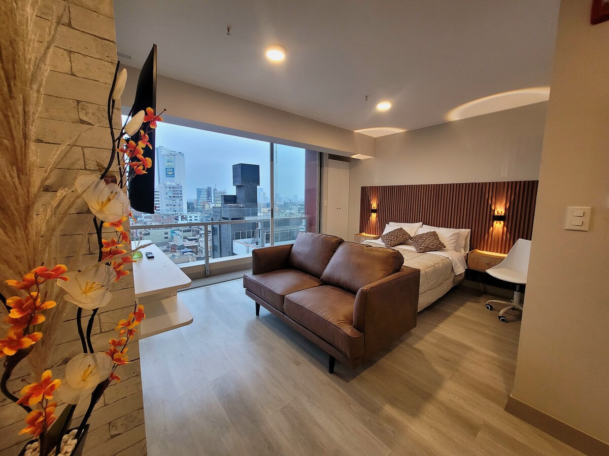 The living area features a brown upholstered sofa and a wall-mounted TV. A spacious bed with neutral bedding is positioned against a wood-paneled accent wall. Large windows provide ample natural light and showcase city views.