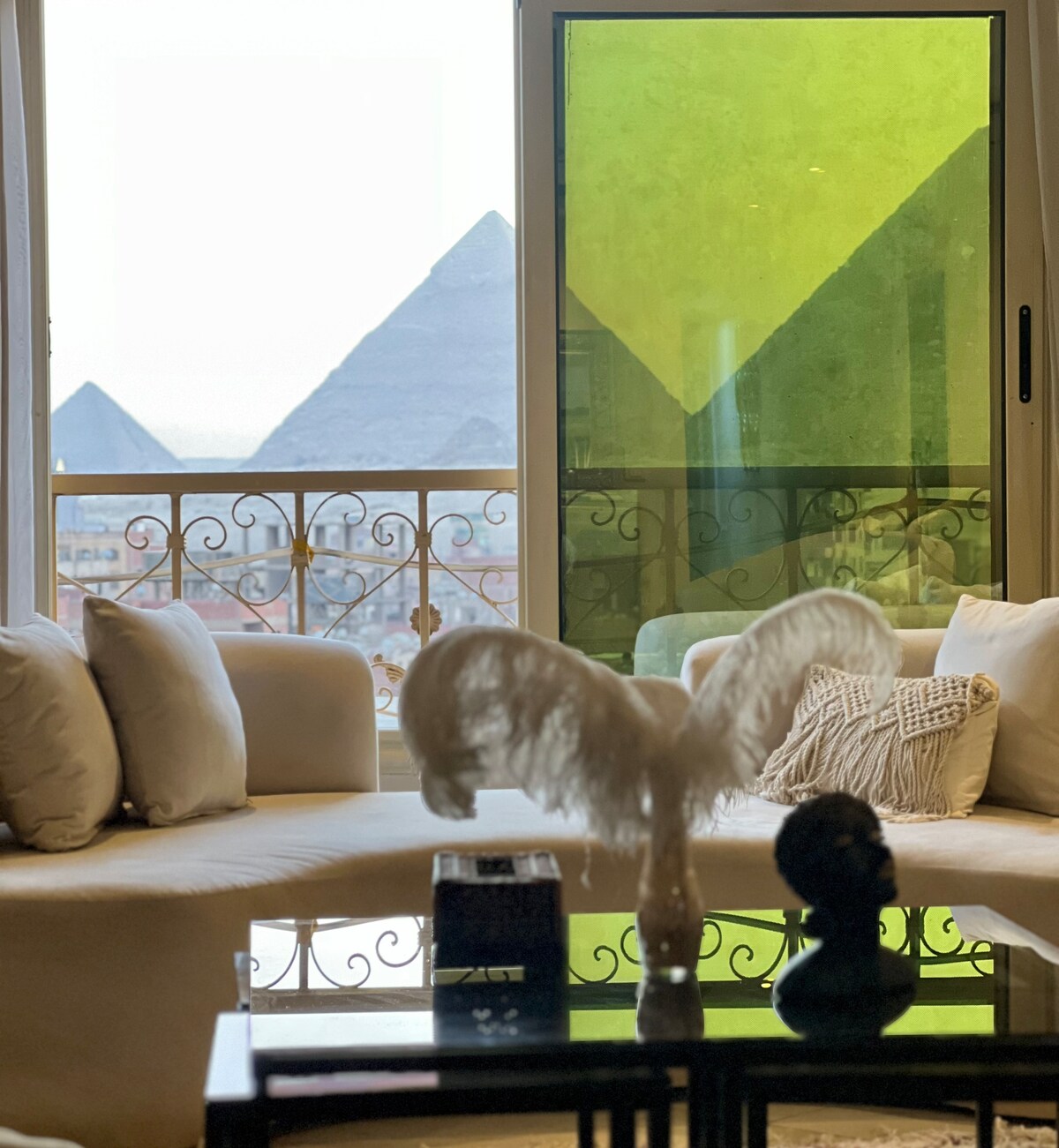The living room features a spacious white sofa adorned with decorative pillows, positioned to enjoy views of the pyramids through large, floor-to-ceiling windows. A modern glass coffee table is set in front, showcasing a small vase and decorative items. Vibrant green glass adds a contemporary touch to the space.