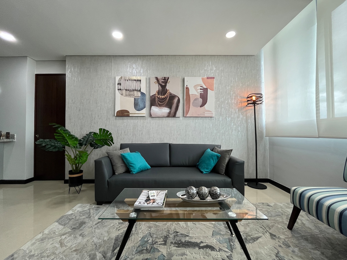 A spacious living area features a modern grey sofa adorned with colorful cushions. A glass coffee table displays decorative items. Artistic wall pieces provide visual interest. Natural light filters through large windows, and a potted plant adds a touch of greenery to the inviting space.
