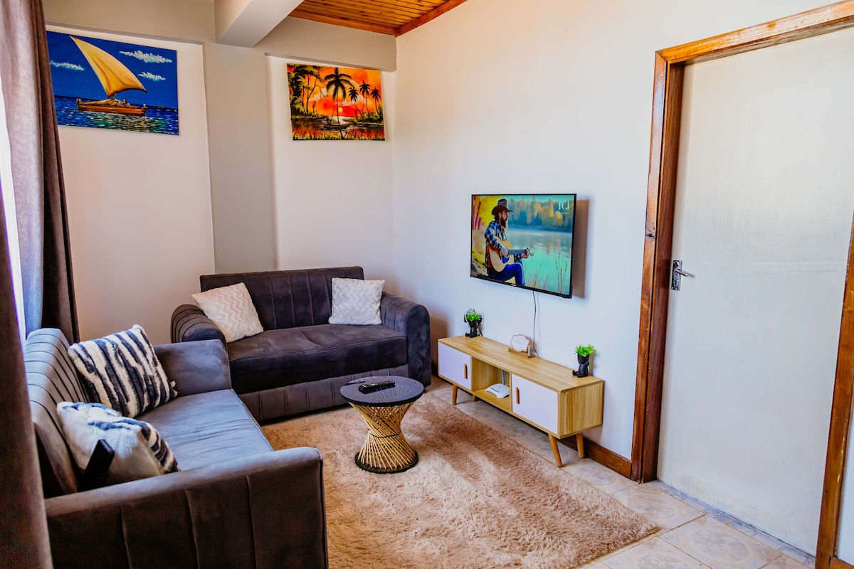 A neatly arranged living area features a gray sofa accompanied by light-colored cushions, sitting on a soft rug. A wooden coffee table sits centrally, while a flat-screen television is mounted on the wall. Colorful artwork depicting landscapes enhances the room's friendly ambiance.