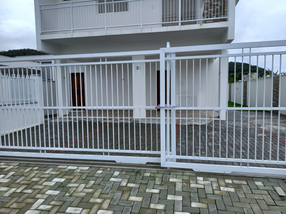 A two-story building is enclosed by a modern white fence. The entrance features a paved area with a rectangular shape leading up to the main door. The exterior walls are light-colored, and there are multiple access points visible, including doors and windows.