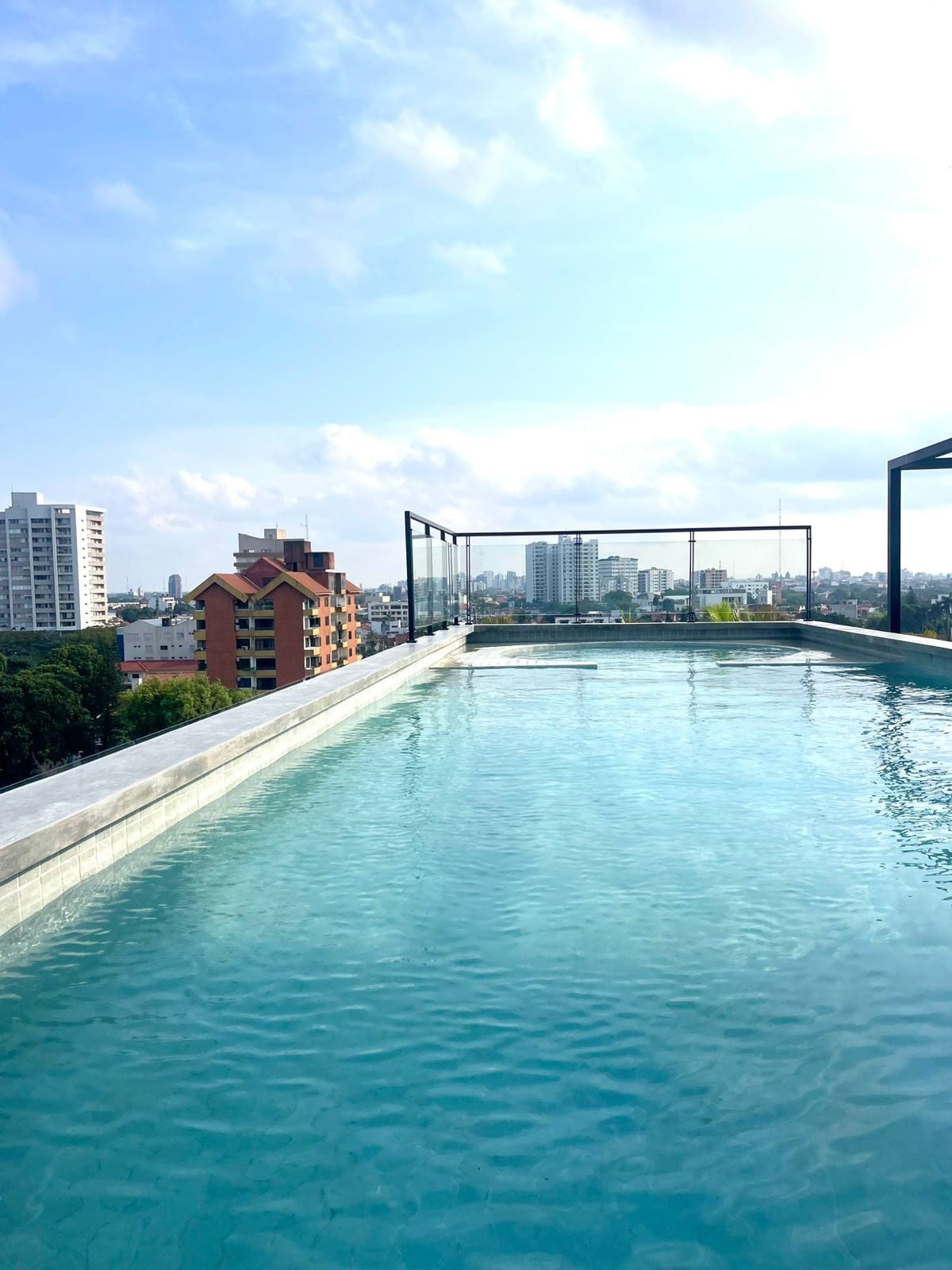 An infinity pool is seen on a rooftop, featuring clear turquoise water that reflects the sky. Surrounding views include various buildings and greenery in the distance, with a bright blue sky visible above.