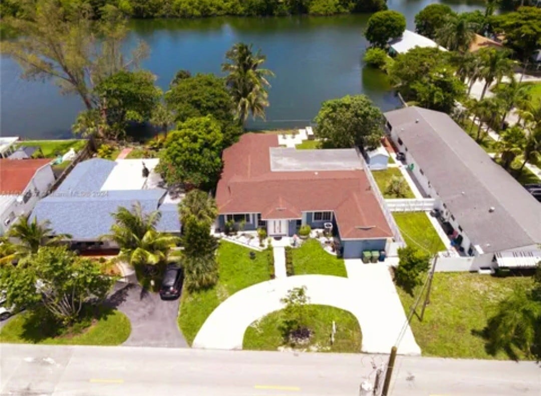 An aerial view captures a spacious home nestled in a lush landscape by a lake. The property features a curved driveway with ample parking space and is surrounded by greenery, creating a serene environment. Neighboring homes can be seen nearby.