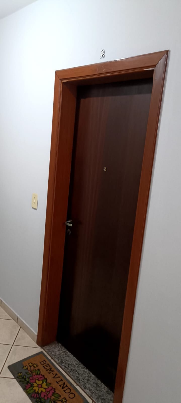 A wooden door with a sleek finish is depicted, featuring a small peephole and a simple handle. The wall surrounding the door is painted in light colors, while a welcome mat with the word 'BEM-VINDO' is placed at the base, adding a friendly touch.