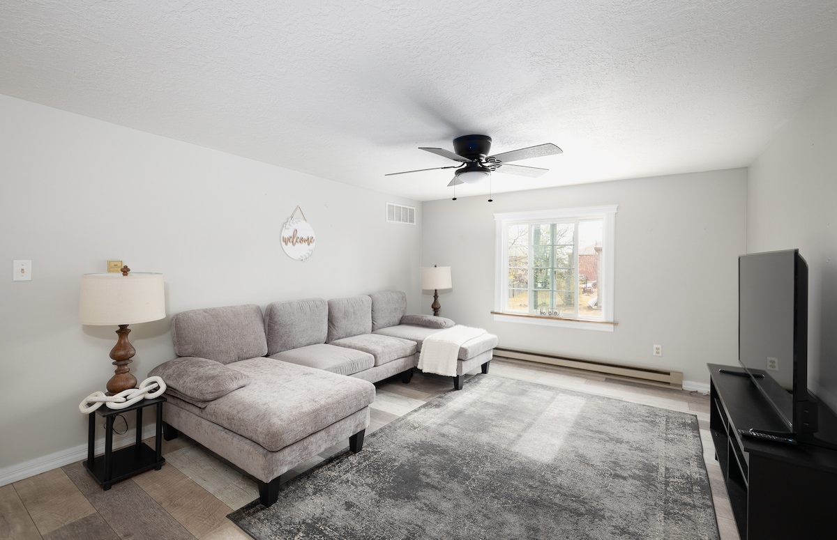 A spacious living room features a large sectional sofa with a light-colored fabric, inviting relaxation. A ceiling fan circulates air, while a flat-screen television is positioned on a dark media stand. Natural light filters through a window, complemented by a decorative lamp on a side table.