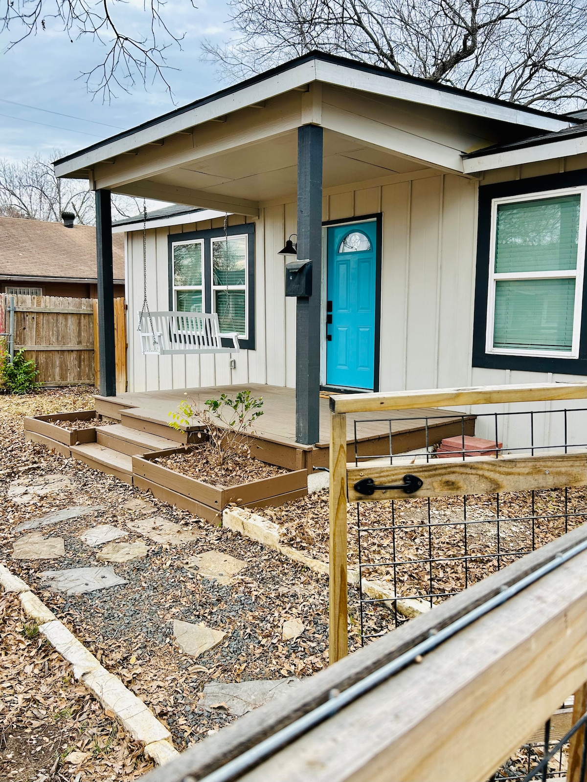 A charming exterior shows a welcoming porch with a bright blue door. Wooden steps lead to the entrance, surrounded by landscaped areas featuring plants and a stone pathway. A wooden fence offers privacy, and trees are visible in the background.