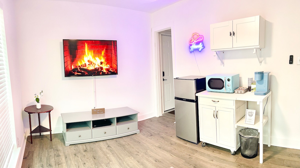 A bright living space is highlighted by a mounted television displaying a fireplace. A small round table sits beside the TV stand. A kitchenette is equipped with a refrigerator, microwave, and coffee maker, alongside clean white cabinetry and a waste bin.