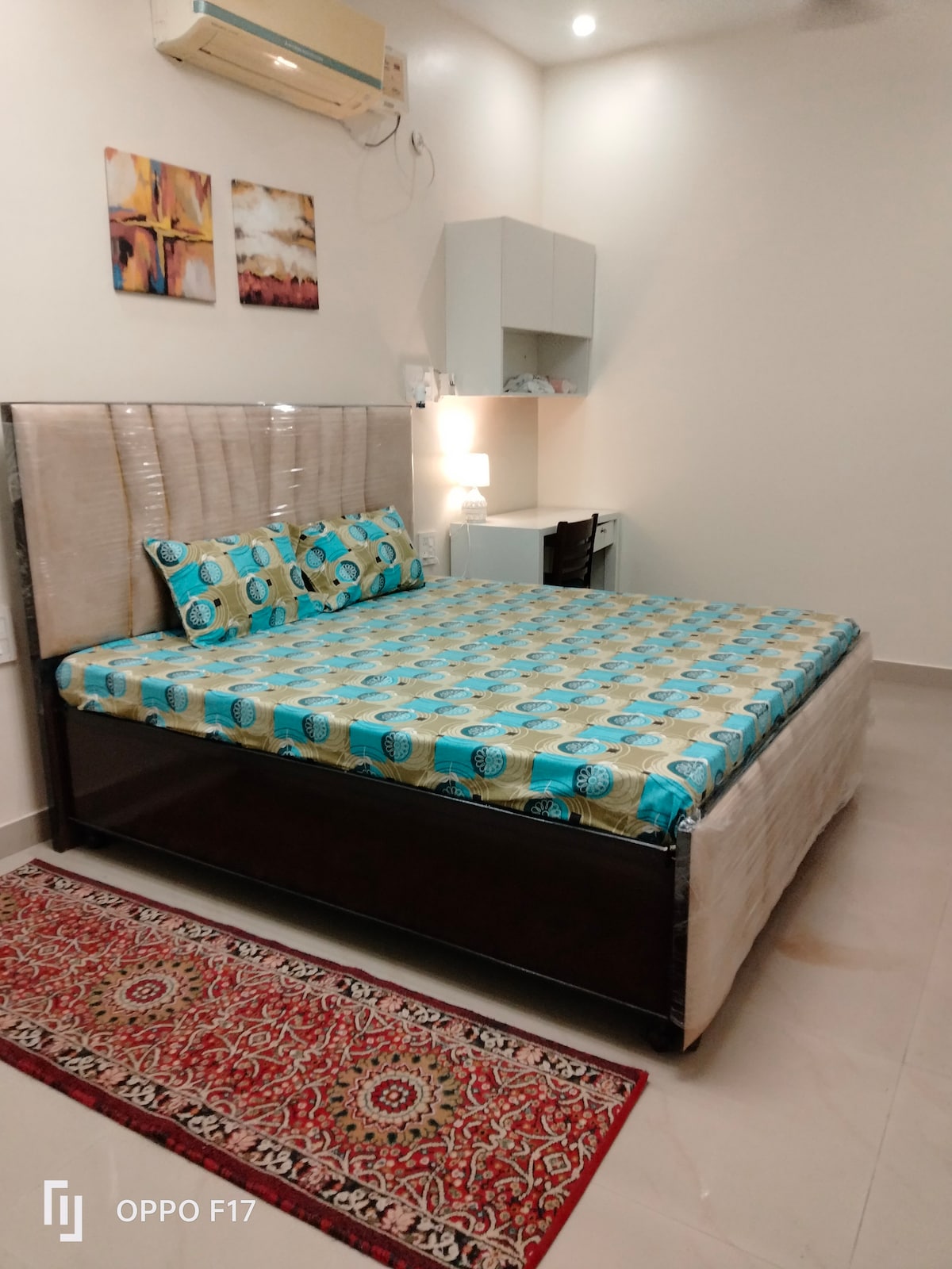 A spacious room is highlighted by a king-size bed adorned with a colorful patterned bedspread. A plush rug adds warmth to the tiled floor. A small desk with a chair and storage cabinets are visible, with soft lighting provided by a bedside lamp.