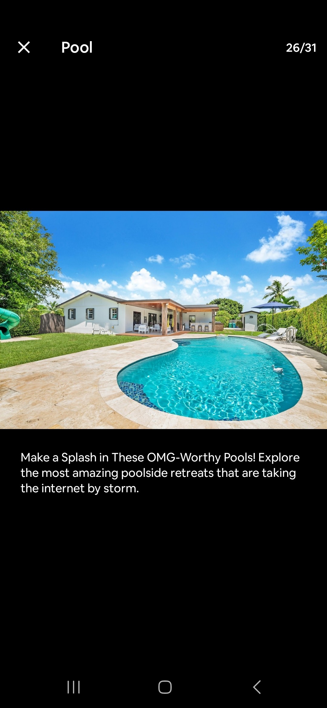 A crystal-clear pool is surrounded by a spacious patio, with lush greenery and palm trees in the background. Bright blue skies and fluffy white clouds create a relaxed atmosphere, while the pool's shape and inviting water highlight an ideal space for leisure.