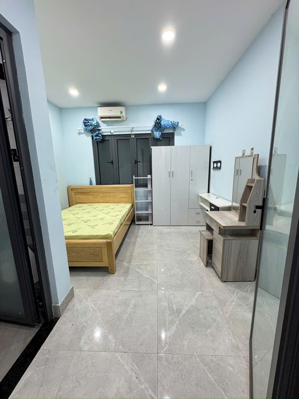 A well-lit bedroom is furnished with a wooden bed and a compact bedside table. A light grey wardrobe and a small desk with a mirror are present. The walls are painted in a soft blue, and a sliding door leads to the outside.