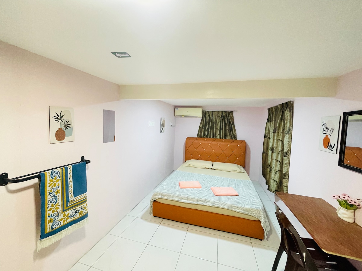 A cozy room features a spacious bed with a brown headboard, accompanied by light-colored bedding. Two orange towels are neatly placed on the bed. A wooden dining table is positioned beside the bed, and plants are showcased in wall art and curtains.