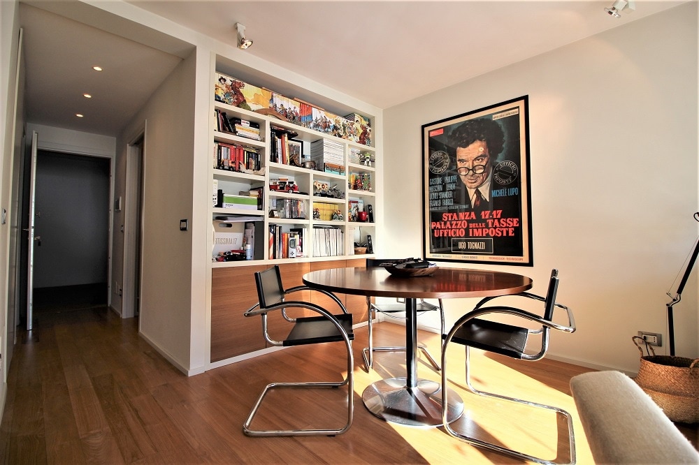 A modern dining area is featured, showcasing a round table surrounded by four sleek chairs. A bookshelf filled with books and decorative items stands against the wall, while natural light illuminates the space through unseen windows, creating an inviting environment.