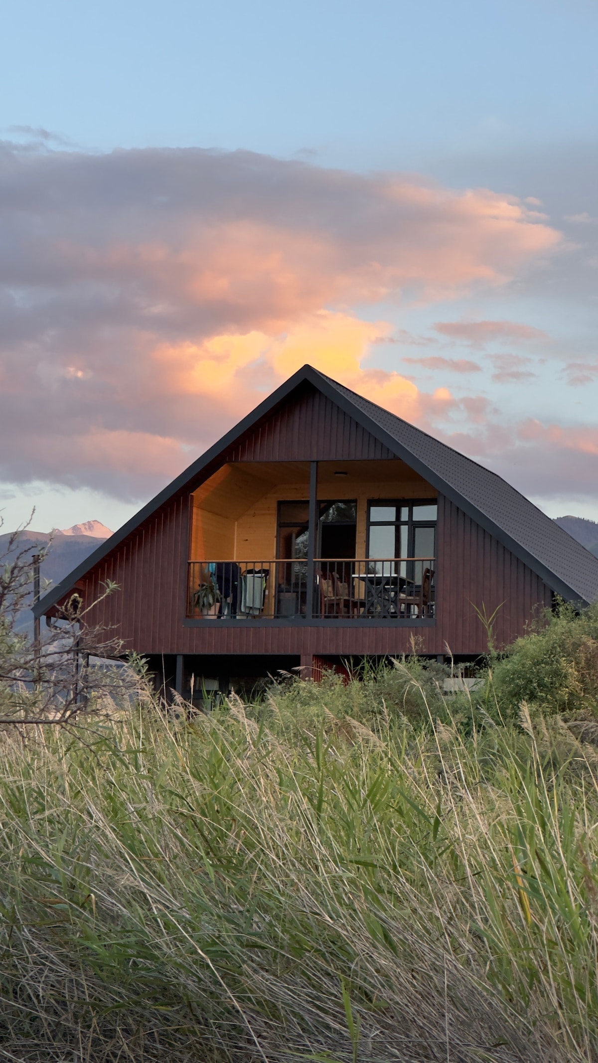 A modern chalet-style cottage presents a striking silhouette against a colorful sky at sunset. Large windows contribute to the structure's aesthetic, while a balcony extends from the upper level, framed by natural grass and vegetation in the foreground.