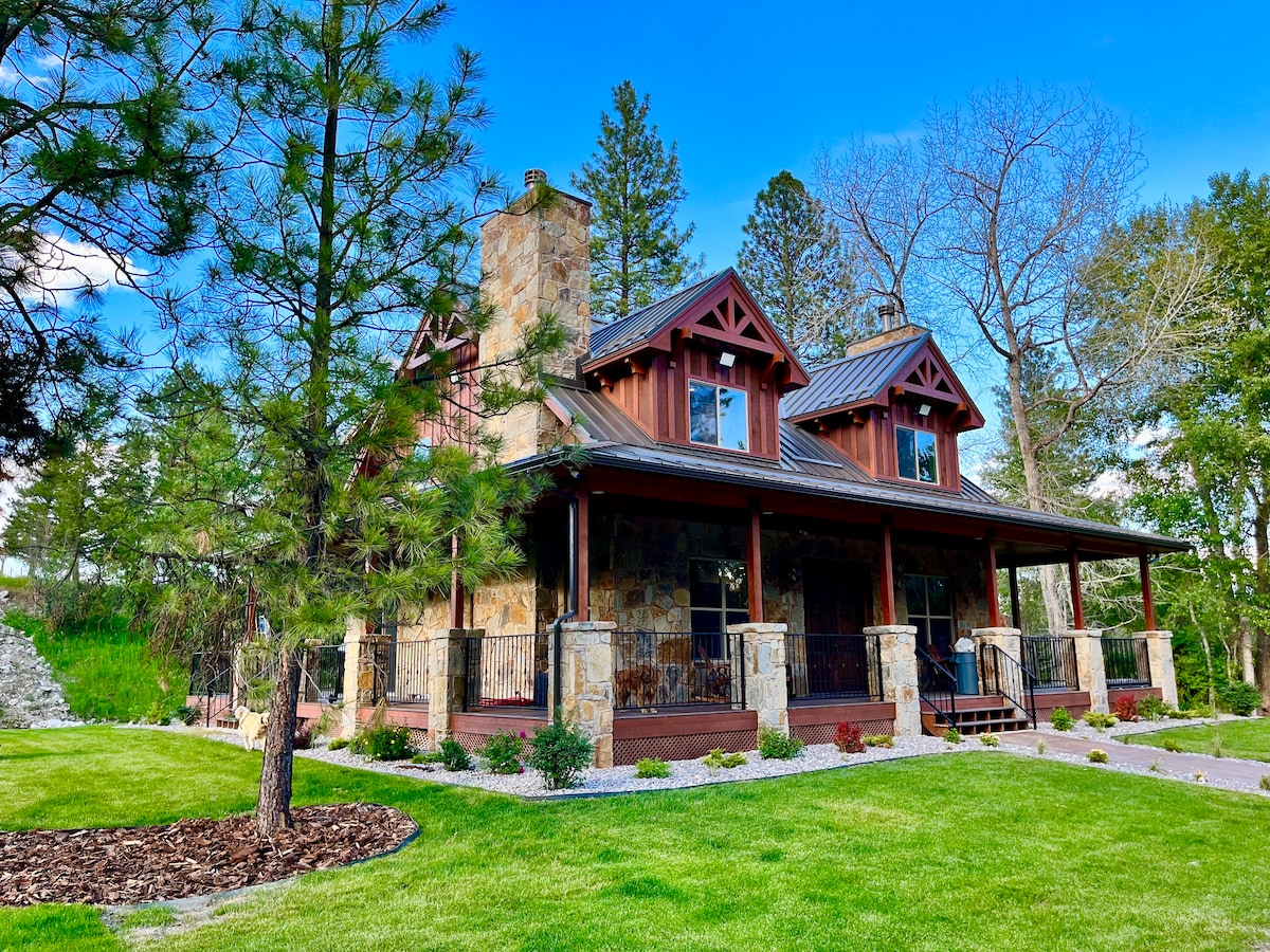 A charming cabin is framed by lush green grass and well-maintained landscaping. The structure features a mix of stone and wood elements, with a covered porch accented by pillars. Pine trees enhance the tranquil outdoor setting, providing a picturesque backdrop.