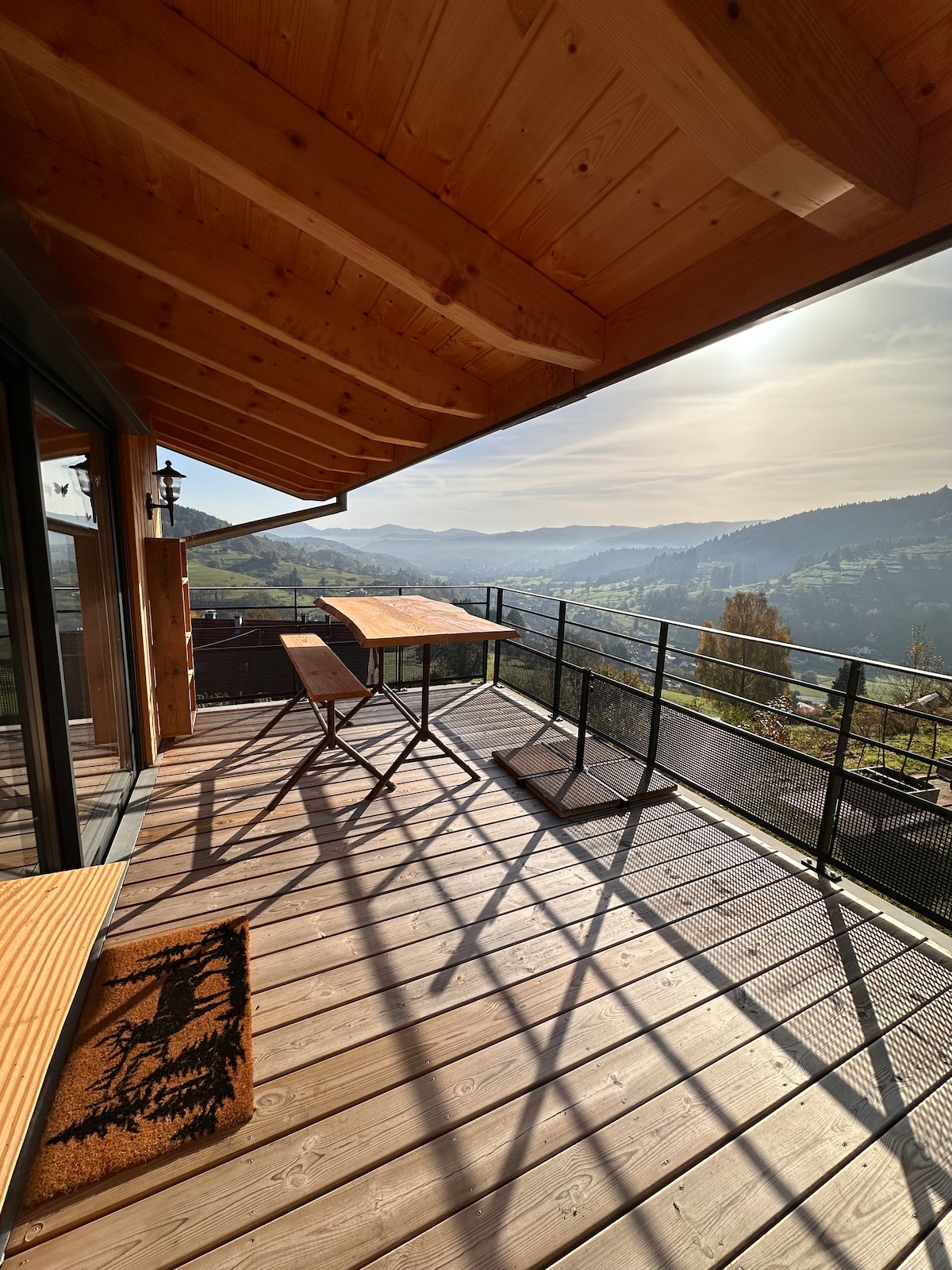 A sunlit balcony features wooden flooring and sleek metal railings, providing expansive views of rolling hills and valleys. Two tables are positioned to catch the sunlight, with shadows creating an intricate pattern on the deck. The natural wood of the roof complements the serene surroundings.