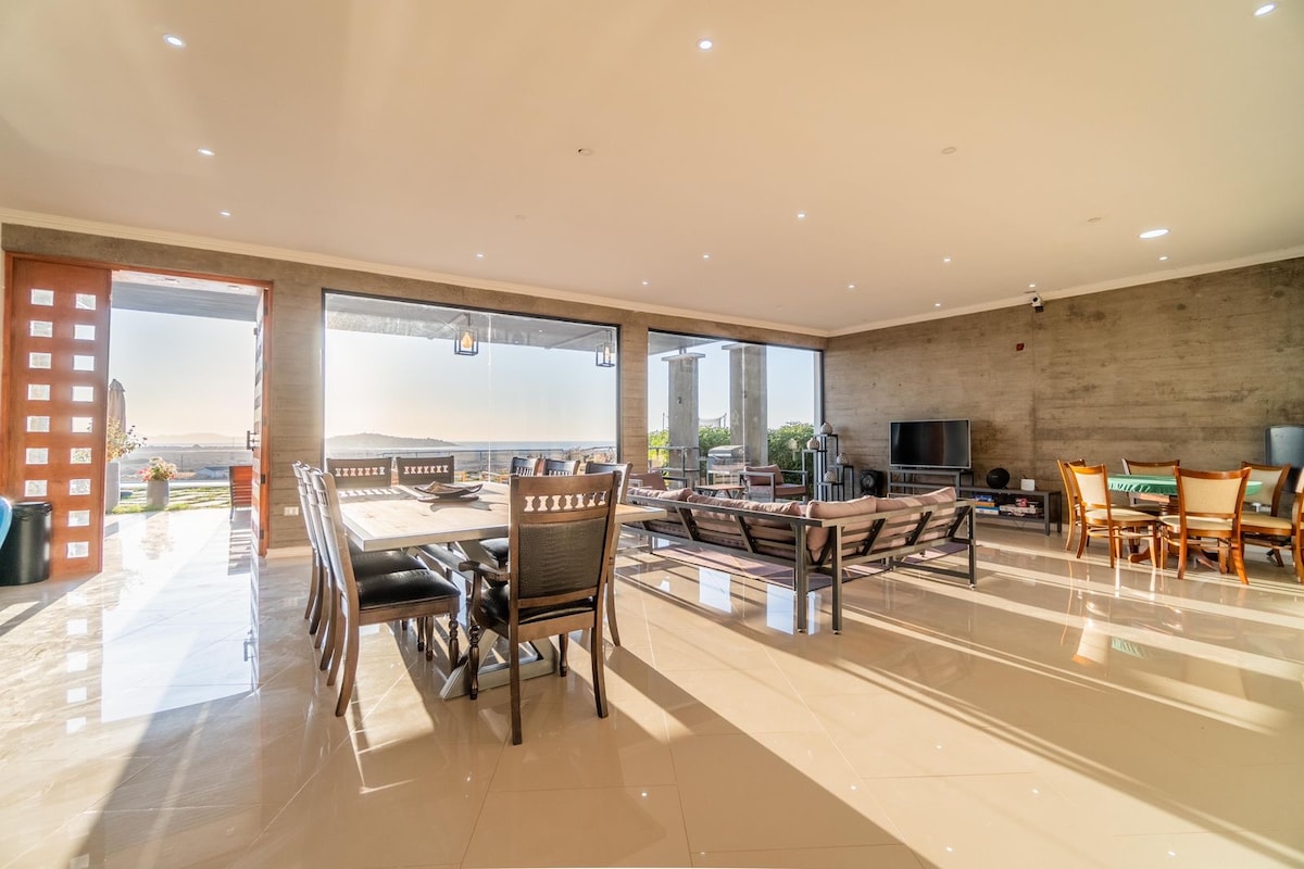 An open-plan living space reveals a large dining table surrounded by comfortable chairs. Natural light filters through expansive glass doors, illuminating the polished floor. Modern furnishings include a stylish sofa and entertainment area, creating a seamless connection between indoor and outdoor areas.