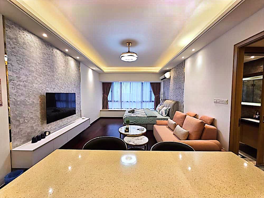 A spacious living area is featured, with a comfortable sofa positioned against the wall and a large bed visible in the background. Natural light filters through sheer curtains, and a flat-screen television is mounted on the wall. A small table is seen in the foreground.