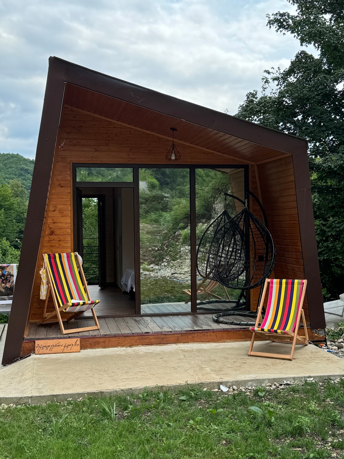 A uniquely designed cabin features a slanted roof and large glass doors that open to nature. Two striped folding chairs sit on the porch, providing a comfortable spot to relax. Surrounding trees create a serene outdoor setting, enhancing the cabin's inviting atmosphere.