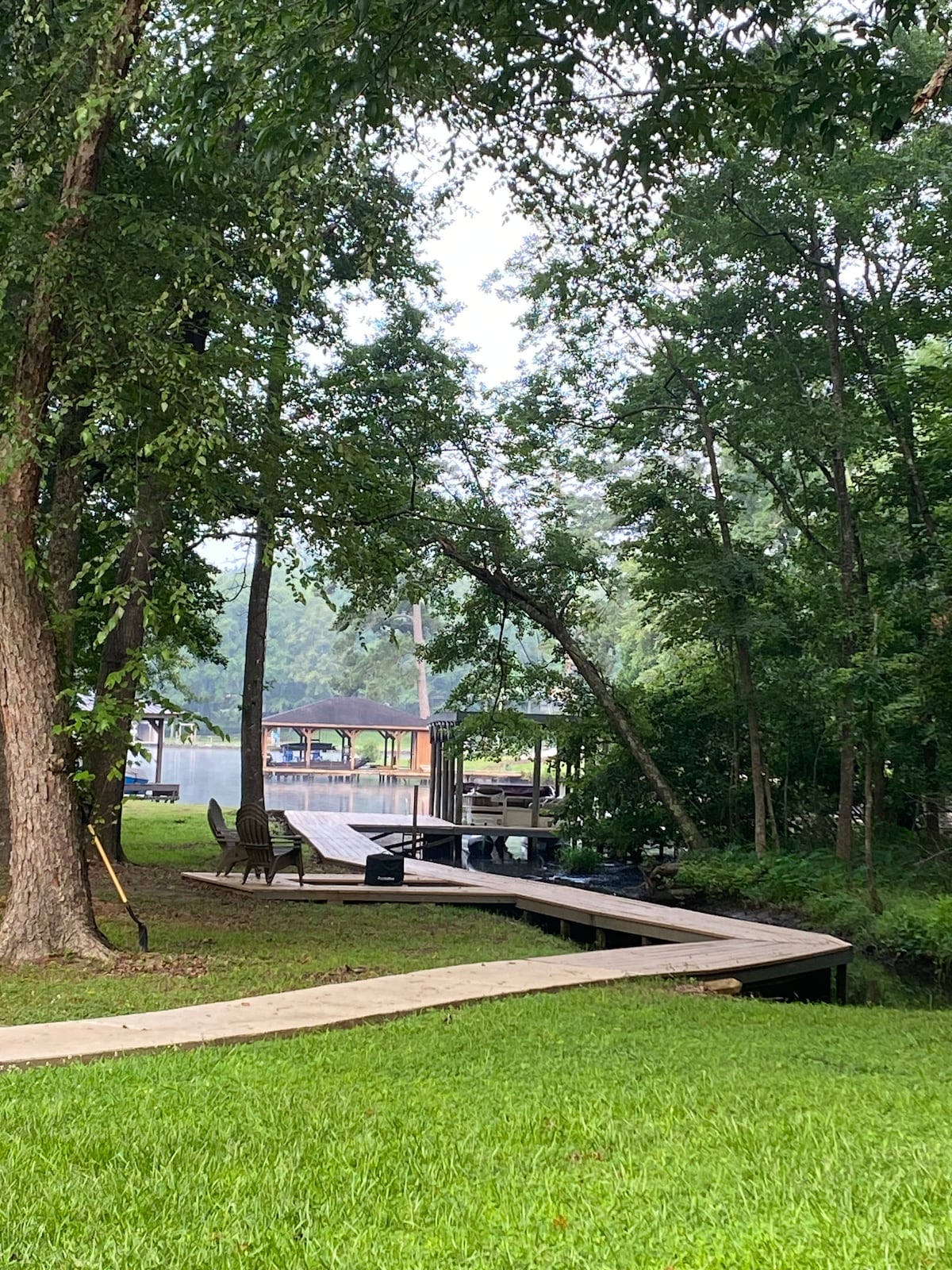 A winding wooden boardwalk is shown extending through lush greenery toward a tranquil lake. Wooden chairs are positioned along the path, surrounded by trees, creating a natural and serene setting. The scene reflects peaceful outdoor access, inviting relaxation by the water.