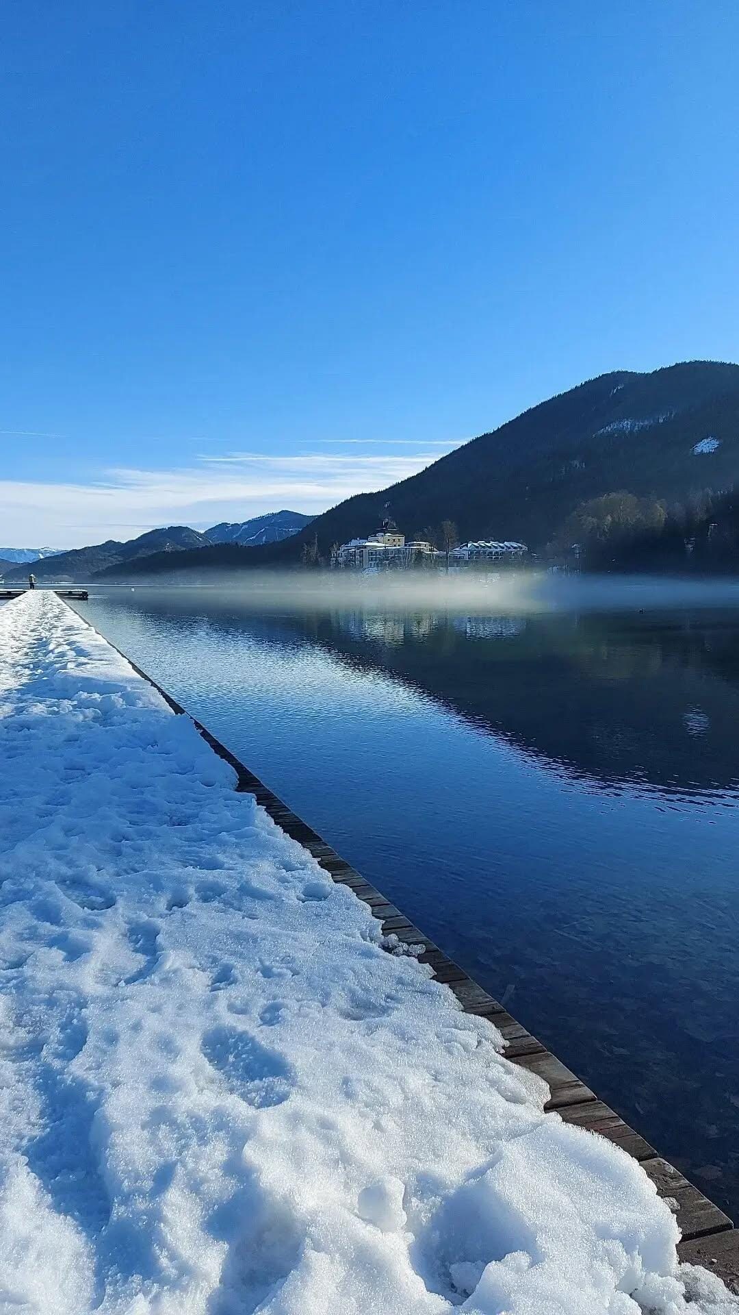 A serene lakeside view showcases a flat wooden dock bordered by fresh snow, reflecting the clear blue sky and surrounding mountains. The still water reveals a peaceful atmosphere, with soft mist rising along the shore, enhancing the tranquil landscape.
