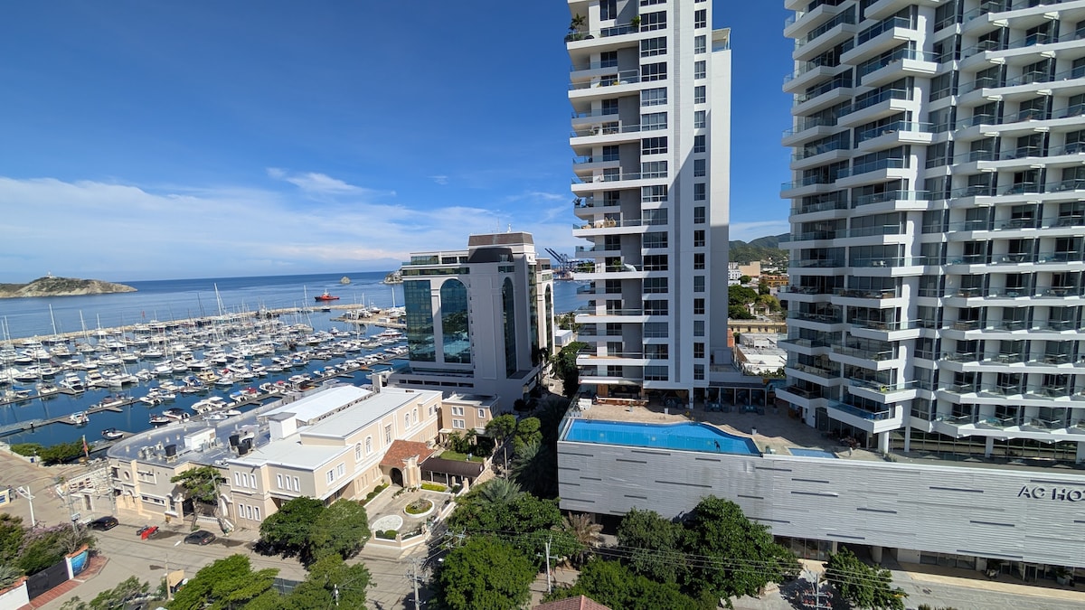A panoramic view showcases modern high-rise buildings in a coastal setting. The marina is filled with boats, while a swimming pool is visible below. Lush greenery surrounds the buildings, complementing the vibrant blue sky and distant ocean views.