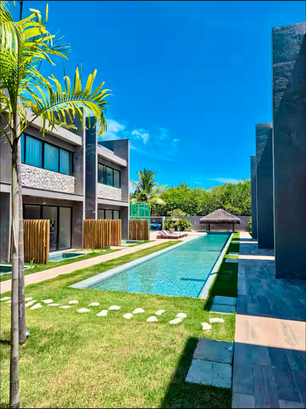 A modern condominium complex is highlighted by an inviting swimming pool that runs alongside well-manicured green lawns. Palm trees and stone pathways accentuate the serene outdoor space, which is complemented by lush greenery in the background and traditional beach huts providing shaded areas.