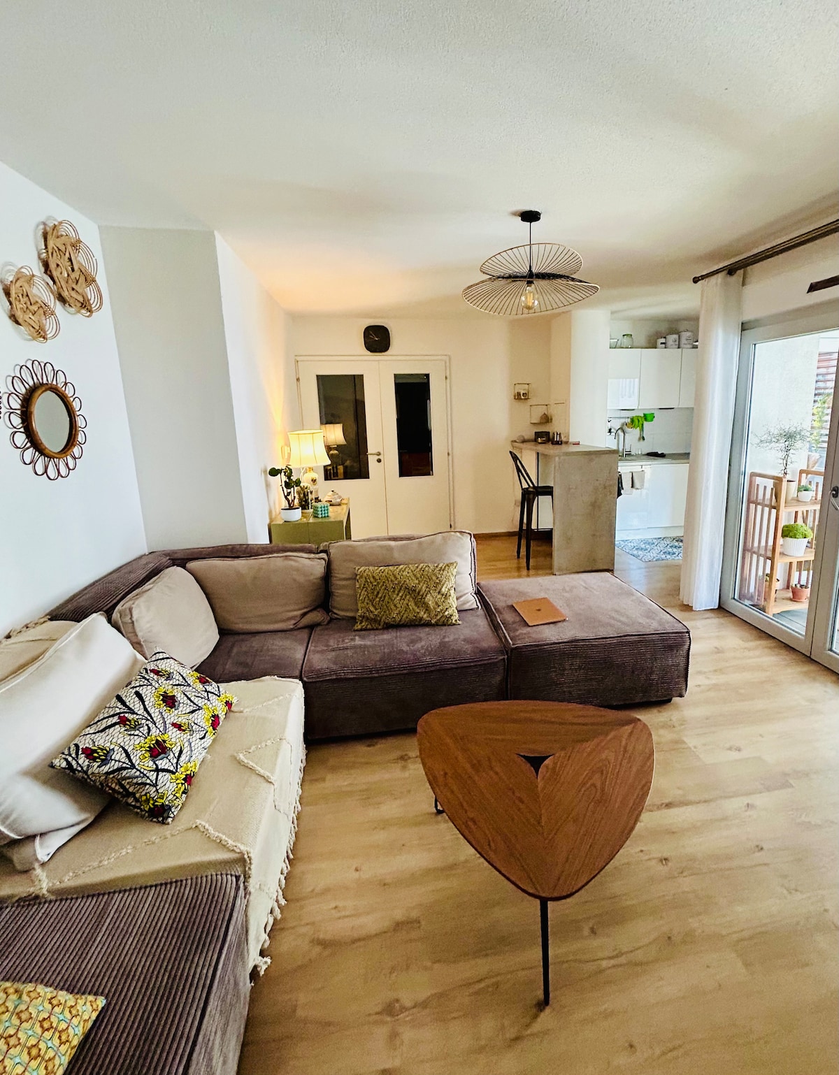 A modern living area is showcased with a sectional sofa adorned with decorative pillows. A unique wooden coffee table is positioned in front, while double doors lead to an outdoor terrace. A dining area is visible in the background, complemented by warm lighting fixtures.