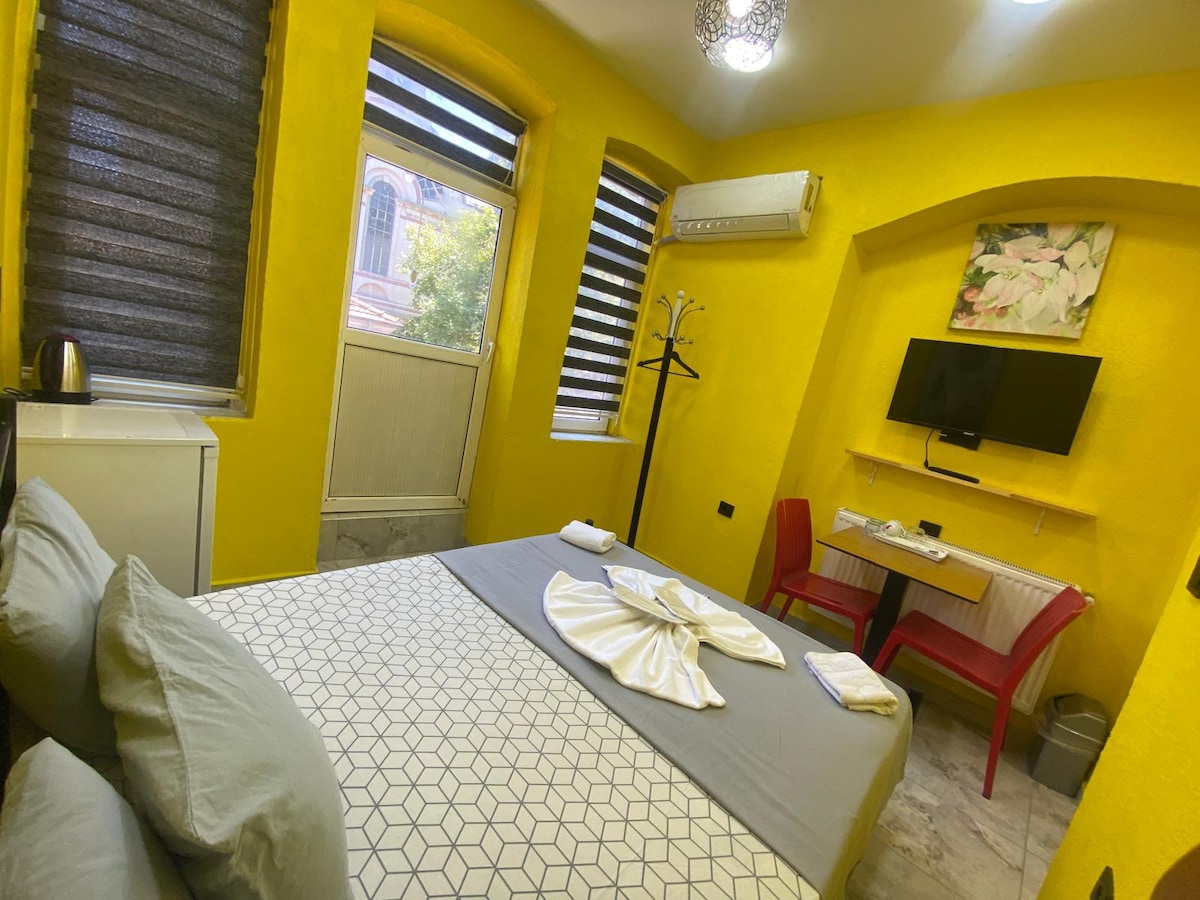 A bright yellow room features a spacious bed with gray linens. A compact refrigerator and kettle are visible. A wall-mounted television is positioned above a table with red chairs. Large windows with horizontal blinds allow natural light to fill the space.