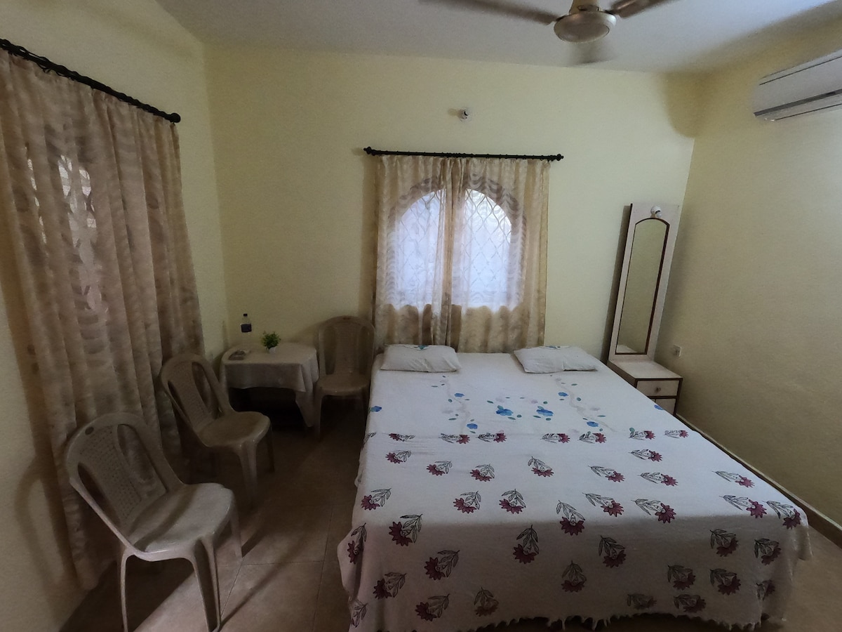 A bright room features a spacious bed with decorative linens and two soft pillows. Light beige walls complement the tiled flooring. Two plastic chairs are placed near a small table, alongside a mirror and a window draped with sheer curtains, allowing natural light to filter in.