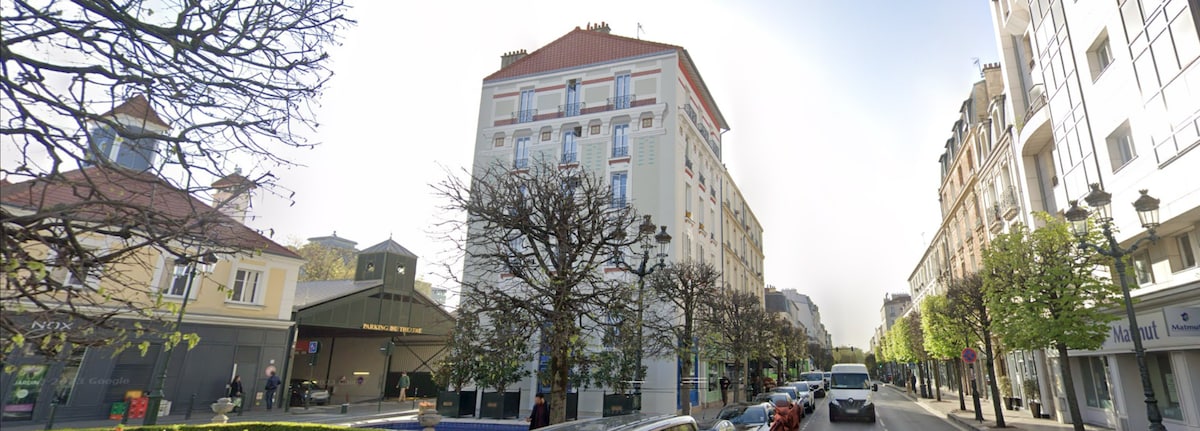 The exterior view of a multi-story building is presented, featuring a combination of classic and modern architectural elements. Large windows line the facade, allowing natural light to fill the interior spaces. Trees with budding leaves are positioned along the street, contributing to the urban setting.