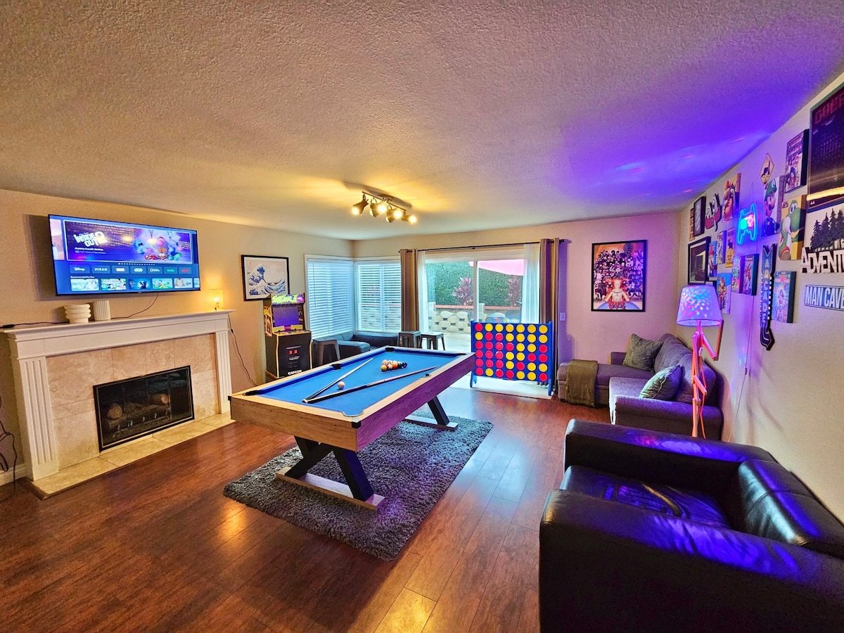 A recreational area features a billiards table centrally located, with a fireplace adorning one side. Large windows allow natural light, illuminating colorful wall art and a vibrant game board. Two leather chairs provide seating, while a cozy rug adds warmth to the space.