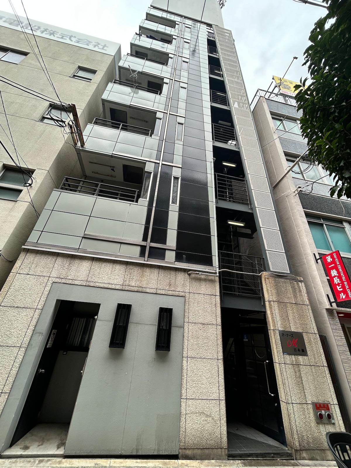 A modern building stands tall with a sleek glass and metal façade. The entrance is flanked by narrow, vertical windows, while the exterior features a blend of textured stone and smooth surfaces, indicating a contemporary design. Surrounding electrical wires add an urban touch.