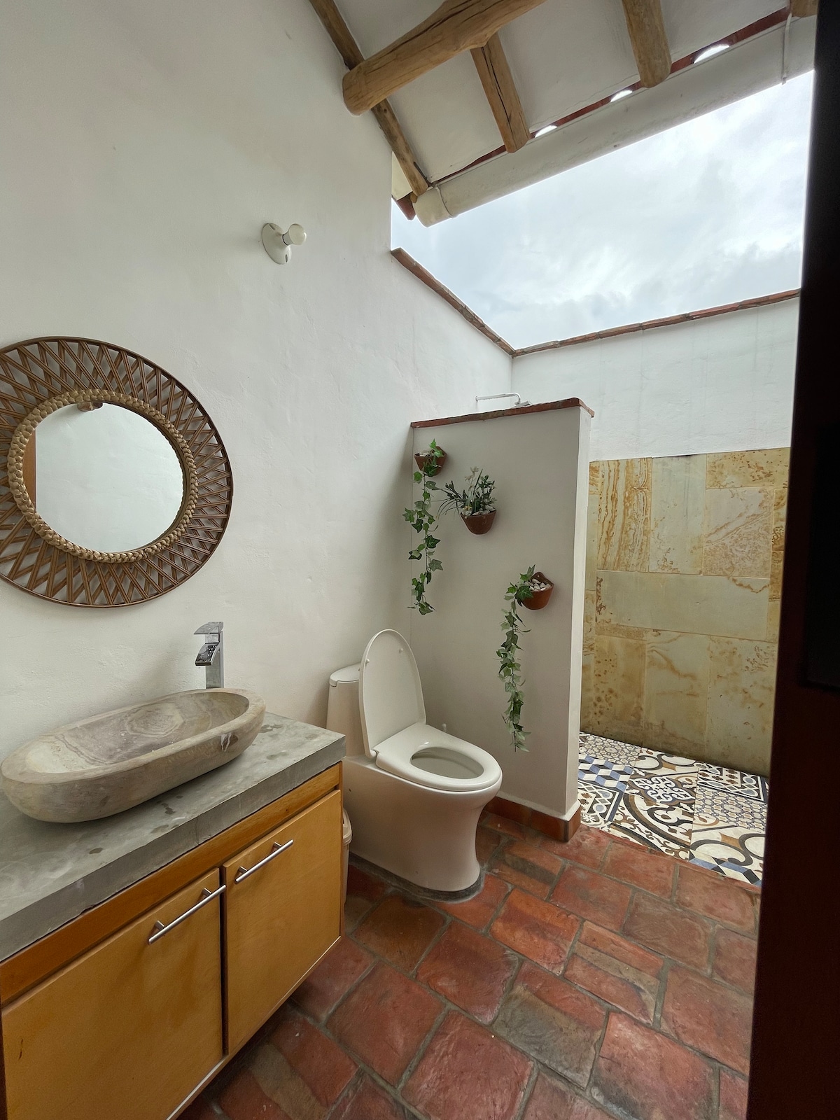 A bright bathroom features a circular mirror above a marble sink. The terracotta floor is complemented by decorative tiles, while an open shower area is adorned with potted plants, offering a natural touch to the space.
