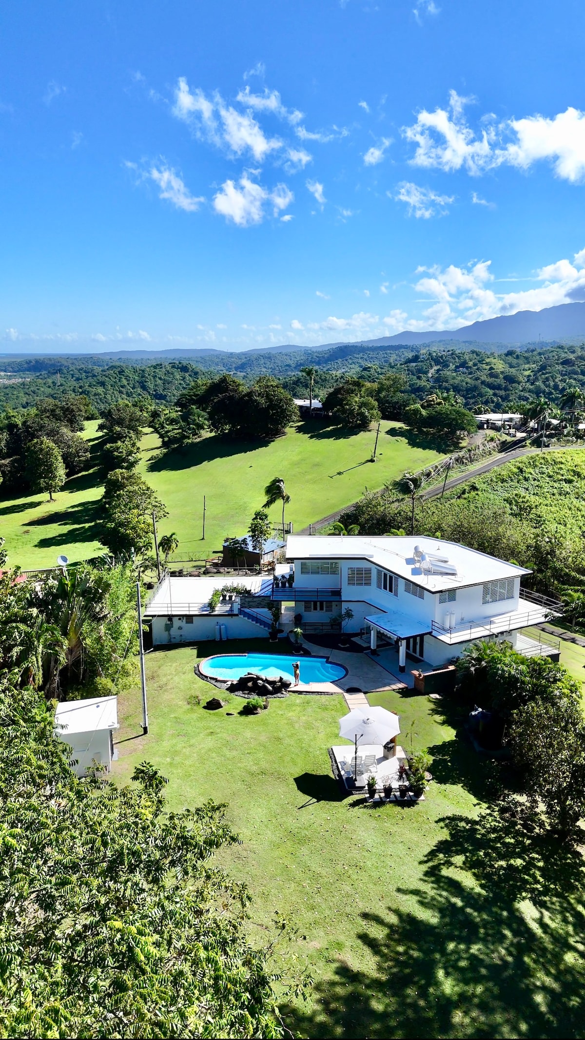 An aerial view showcases a modern hacienda set on a spacious lawn surrounded by lush greenery and mountains. The property features a turquoise pool and a shaded outdoor seating area, all under a clear blue sky adorned with scattered clouds.
