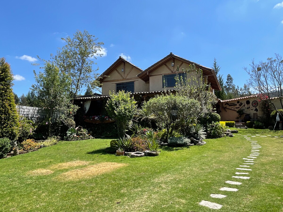 A serene garden is featured, with a variety of plants and trees surrounding a spacious lawn. The rustic two-story house, with a terracotta roof, is set against a clear blue sky, providing a tranquil backdrop for relaxation.